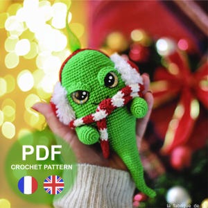 May include: Green crochet Grinch-like character wearing a red and white striped scarf and white earmuffs. The character has large black eyes and a grumpy expression.  PDF Crochet Pattern.