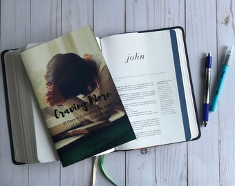 May include: A closed book titled "Craving More: 21 Days to Cultivating the Habit of Studying God's Word" by Michelle Trabon, with a white ribbon bookmark, is open to the book of John in a Bible.