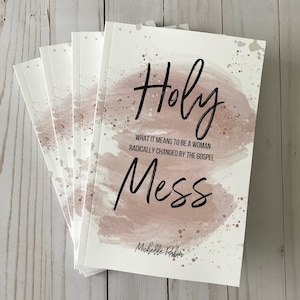May include: A stack of books with a pink watercolor background and the title "Holy Mess" in black lettering. The subtitle reads "What it means to be a woman radically changed by the gospel." The author's name is "Michelle Raben".