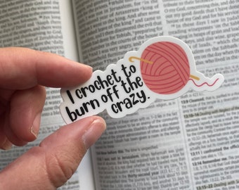 May include: A white sticker with the text "I crochet to burn off the crazy" and a pink yarn ball with a yellow crochet hook. The sticker is held in front of an open book.