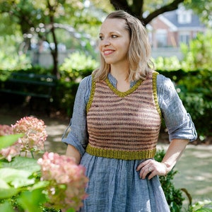 Può includere: Una donna indossa un gilet di maglia a righe con un bordo verde sopra un vestito di lino blu. È in piedi in un giardino con vegetazione verde e fiori rosa.