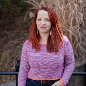 May include: A cropped, long-sleeved sweater in a textured lavender knit with coral trim. The sweater is paired with dark wash jeans and a brown belt. The model has red hair and is wearing earrings.