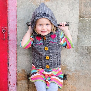 May include: A young girl wearing a gray hooded vest with colorful hearts, a striped shirt, gray leggings, and silver boots. She is sitting on a brick wall.