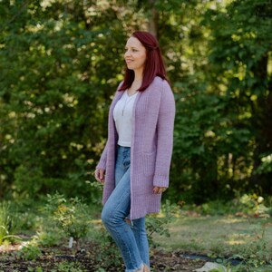 May include: A woman wearing a light purple cardigan over a white t-shirt and blue jeans. She is standing in a garden with green foliage and purple flowers.