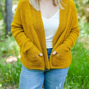 Può includere: Una donna indossa un cardigan giallo senape con tasche sopra una camicia bianca a collo a V e jeans blu con dettagli strappati.