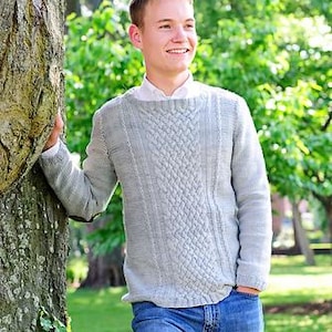 Könnte beinhalten: Hellgrauer Strickpullover mit Zopfmuster auf der Vorderseite. Der Pullover hat lange Ärmel und wird mit einer blauen Jeans kombiniert. Die Person lehnt an einem Baum in einem Park.