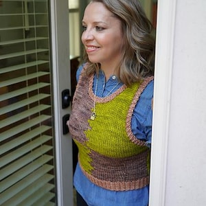 Könnte beinhalten: Eine gestrickte Weste in Grün-, Braun- und Beigetönen. Die Weste wird über einem blauen Jeanshemd getragen. Die Weste hat ein einzigartiges Farbblock-Design und einen gerippten Saum. Die Person steht in einem Türrahmen.