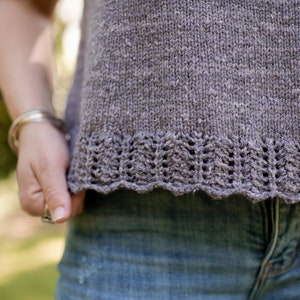 May include: A close-up of a gray knitted top with a lacy trim. The top is worn by a person and the bottom of the top is tucked into blue jeans.