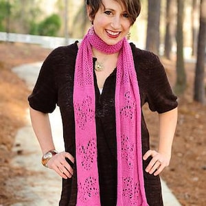 May include: A woman wearing a dark brown short-sleeved top and blue jeans. A bright pink, open-weave scarf is draped around the neck. The scarf has a delicate, lacy pattern. A gold-toned pendant necklace and a bracelet are also visible.