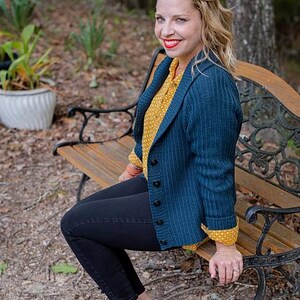 Può includere: Una donna indossa un cardigan blu scuro sopra una camicetta gialla, pantaloni neri e ballerine blu. Il cardigan ha una chiusura con bottoni e una trama a coste. La donna è seduta su una panchina di legno.
