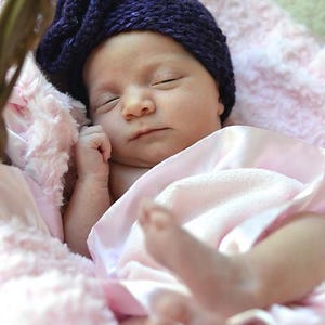May include: A newborn baby wearing a dark blue knitted cap with a flower design, resting peacefully. The baby is wrapped in a soft pink blanket and resting in a pink fluffy basket. The image is a close-up shot.
