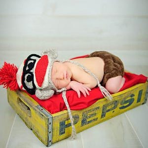 May include: A newborn baby wearing a monkey hat with a red pom-pom and brown diaper cover, lying on a red cushion inside a yellow Pepsi crate. The crate has black lettering.