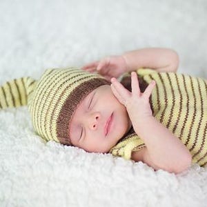 May include: A newborn wearing a yellow and brown striped knit hat and matching outfit. The baby is lying on a white, fluffy surface, with eyes closed and hands near the face. The outfit includes a long-sleeved top and pants.