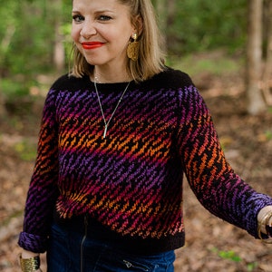 Può includere: Una donna indossa un maglione nero e viola con un motivo diagonale di arancione, rosso e giallo. Indossa anche pantaloncini di jeans e gioielli d'oro.