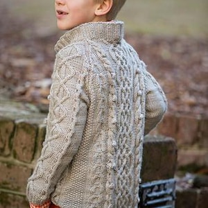 May include: A light gray cable-knit sweater with a high collar. The sweater features intricate patterns and textures, with a mix of cable and bobble stitches. The sleeves are long, and the sweater is worn with orange pants.