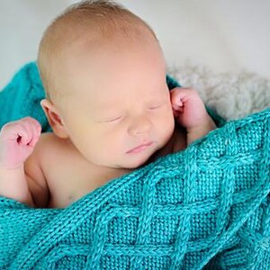 May include: A newborn baby wrapped in a teal knitted blanket. The baby is sleeping peacefully, with closed eyes and tiny hands near its face. The blanket has a textured, cable-knit design, creating a cozy and comforting scene. The background is a soft, neutral color.