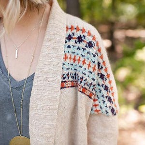 Pode incluir: Cardigã de malha bege com um padrão geométrico colorido na manga superior. O cardigã é combinado com uma camiseta cinza com decote em V e colares dourados em camadas. O fundo está desfocado com folhagem verde.