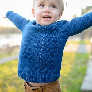 Puede incluir: Un suéter azul tejido con un diseño de punto trenzado en la parte delantera. El suéter tiene mangas largas y un dobladillo acanalado. El niño lleva pantalones marrones. El fondo está borroso, con hierba verde y árboles visibles.
