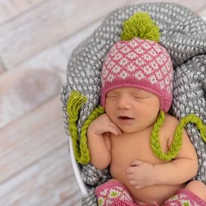 May include: A newborn baby wearing a pink and white patterned hat with a green pom-pom and matching booties. The baby is nestled in a gray blanket inside a white bowl. The hat has green braided ties.