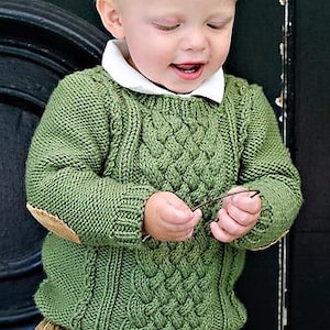 May include: A green cable-knit sweater with elbow patches and a white collared shirt. The sweater has a textured design and is worn with khaki pants. The child is holding a small twig.
