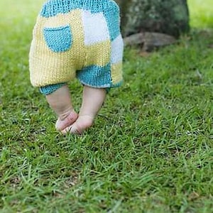 Puede incluir: Unos pantalones cortos de bebé tejidos con un diseño de patchwork. Presentan cuadrados azules, amarillos y blancos. Se ve un pequeño bolsillo en la parte trasera. El bebé está sobre hierba verde.
