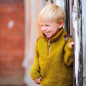 May include: A child in a mustard yellow knitted jumper with a zip and brown leather shoulder patches, paired with blue jeans. The child smiles near a weathered wooden door. The sweater's texture is visible.