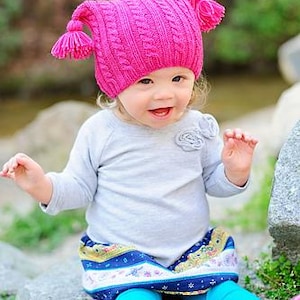 May include: A child in a vibrant pink knitted hat with ear flaps and tassels. The child is also wearing a light grey long-sleeved top, a patterned skirt with blue and gold details, and turquoise leggings. Pink shoes complete the outfit.