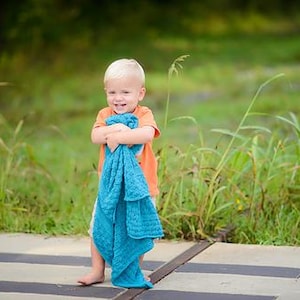 May include: A young child wearing an orange shirt and holding a turquoise blue towel. The child is standing on a concrete surface with black stripes, with a green grassy background. The child is smiling.