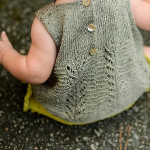 Puede incluir: Vestido de bebé tejido en un suave color verde oliva. El vestido sin mangas presenta una espalda abotonada con tres botones de madera. La parte inferior del vestido tiene un forro amarillo brillante. El vestido está diseñado con un delicado calado.