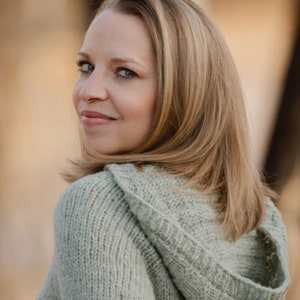 Può includere: Una donna con lunghi capelli biondi indossa un maglione con cappuccio verde chiaro. Il maglione è lavorato a maglia e ha una vestibilità ampia.