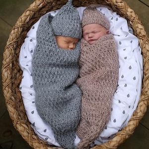 May include: Two newborn babies, each in a knitted cocoon and hat, resting in a woven basket. One is wrapped in grey, the other in a soft brown. The basket is lined with white fabric featuring a grey raindrop pattern.