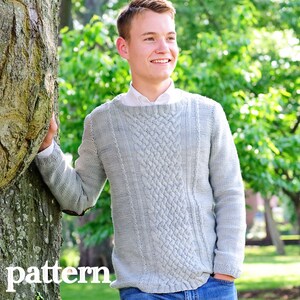 Puede incluir: Un suéter de punto gris claro con un patrón de trenza en la parte delantera. El suéter tiene cuello redondo y mangas largas.