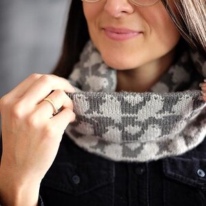 Könnte beinhalten: Ein grau-weiß gemusterter Rundschal. Der Schal ist aus weichem Strickmaterial gefertigt und weist ein sich wiederholendes geometrisches Design auf. Die Person im Bild trägt ein dunkelblaues Jeanshemd.