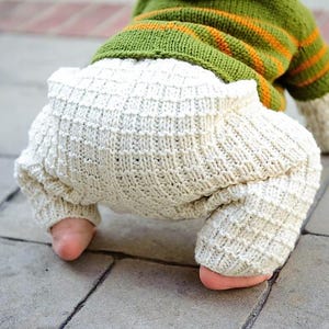 May include: A close-up of a baby wearing a knitted outfit. The outfit features white, textured bloomers and a green and orange striped top. The baby's feet are visible, adding a sense of scale and detail to the image.