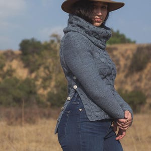 Può includere: Un maglione blu a maglia con collo alto e bottoni laterali. Il maglione ha un motivo a treccia e viene indossato con jeans blu e un cappello marrone.