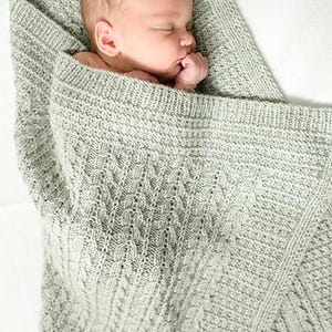 Könnte beinhalten: Ein schlafendes Baby, eingehüllt in eine weiche, salbeigrüne Strickdecke. Die Decke hat ein strukturiertes Zopfmuster und einen gerippten Rand. Das Baby liegt bequem, das Gesicht ist sichtbar, und die Hand ist in der Nähe des Mundes.