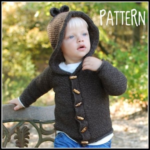 Puede incluir: Una chaqueta de punto marrón con capucha y botones de madera para un niño pequeño. La chaqueta tiene una capucha con orejas de oso y está hecha de punto texturizado.