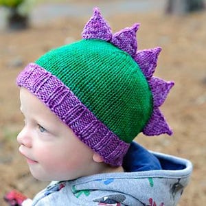 Peut inclure: Un bonnet de dinosaure tricoté vert et violet. Le bonnet présente une couronne verte avec des pointes violettes et un bord violet. Le chapeau est conçu pour ressembler à un dinosaure. Le chapeau est en tricot doux.