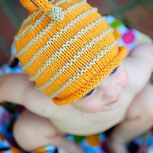 May include: A knitted baby hat in bright orange with horizontal tan stripes. The hat has a small loop and a tan ball on top. The baby is sitting on a colorful polka dot blanket.
