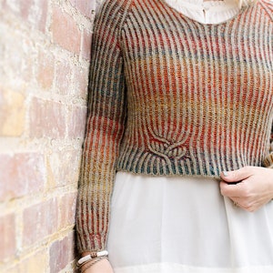 May include: A striped, multi-coloured sweater with a leaf design on the front. The sweater is cropped and has long sleeves.
