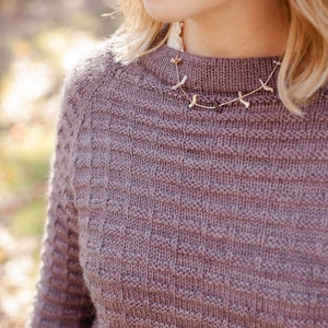 Può includere: Primo piano di una donna che indossa un maglione di maglia color malva con una delicata collana d'oro con una serie di piccoli ciondoli a forma di uccello.