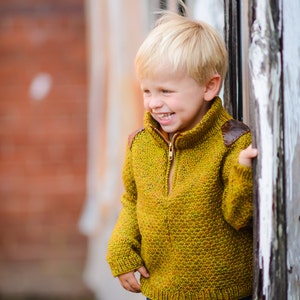 May include: A young boy with blond hair smiles while wearing a yellow and brown jumper and blue jeans. He is standing next to a weathered wooden wall.