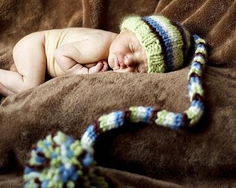 May include: A newborn baby wearing a green, blue, and white striped knit hat with a long, decorative tail, resting on a brown blanket. The baby is curled up, sleeping peacefully.