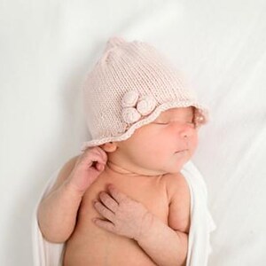 May include: A newborn baby wearing a light pink knitted hat with three decorative flower-shaped accents. The baby is lying on a white surface, wrapped in a white cloth.