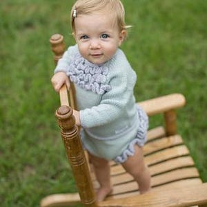 May include: A baby wearing a light blue knitted outfit with a ruffled collar and matching diaper cover, standing on a wooden rocking chair. The baby has blonde hair and is looking at the camera. The background is green grass.