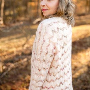 Könnte beinhalten: Eine cremefarbene Strickjacke mit Zickzackmuster. Der Pullover hat eine lockere Passform und verfügt über dezente rosa und goldfarbene Akzente im gesamten Design. Das Model trägt den Pullover im Freien.