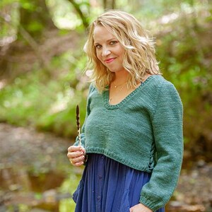 May include: A woman wearing a teal green cropped sweater with long sleeves and a V-neck. The sweater is paired with a long, navy blue dress. She is holding a brown feather. The background is a blurred forest scene.