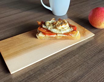 May include: A wooden serving board with a light brown gradient to a white color. A small sandwich with a fried egg, bacon, tomato, and cheese is on the board. A red apple is on the table next to the board.