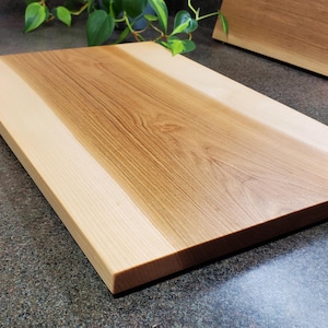 May include: A rectangular wooden cutting board with a light wood border and a darker wood center. The board is made of two different types of wood, creating a unique and beautiful design.