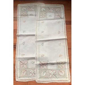 May include: Two cream-coloured rectangular table runners with leaf embroidery in green, red, and brown. The edges have a white crocheted trim. The design features leaf patterns and small scattered leaf motifs.